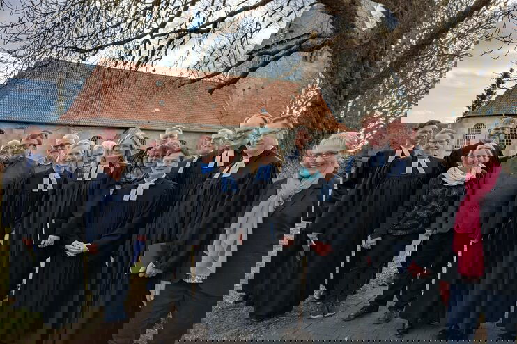 Gut ein Dutzend Personen, teils in Talaren, stehen unter einem Baum vor einer Kirche.