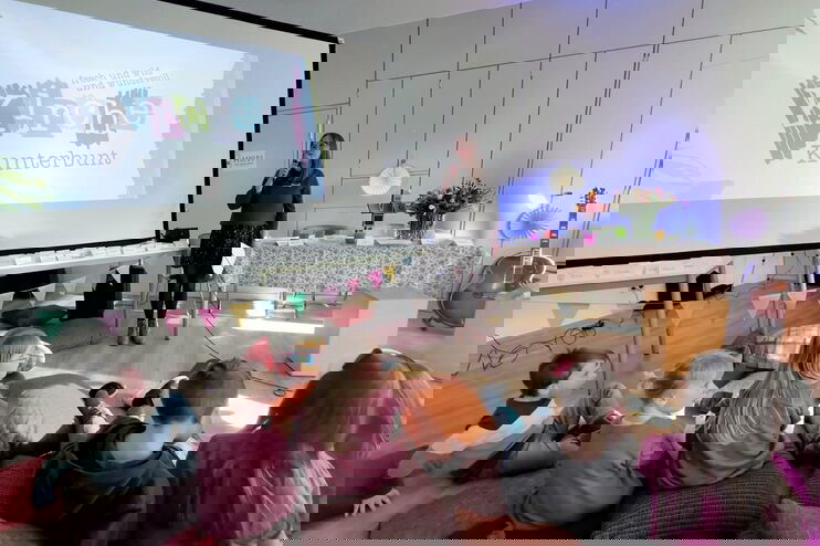 In einem Raum steht an einer Leinwand "Kirche kunterbunt", daneben eine weiblich gelesene Person mit einem Mikro. Vor ihr sitzen Kinder auf vielen bunten Kissen.