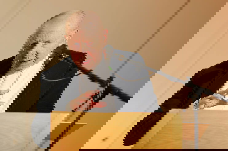 Eine ältere als Mann lesbare Person steht an einem Rednerpult und spricht in ein Mikrofon.