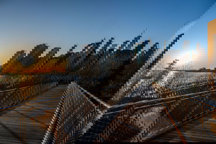 Blick auf Hochhäuser vor strahlend blauem Himmel von einer Brücke aus. Links geht die Sonne über Wasser unter.