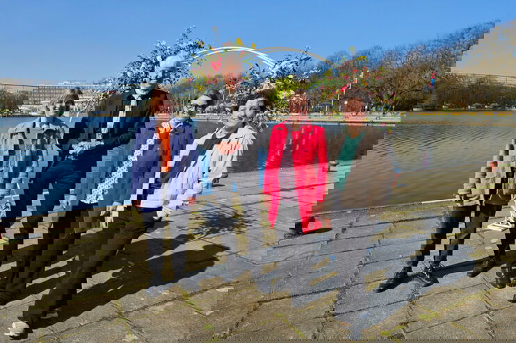 Eine Gruppe steht vor einem See. Drei davon sind als Frauen lesbar, einer als Mann.