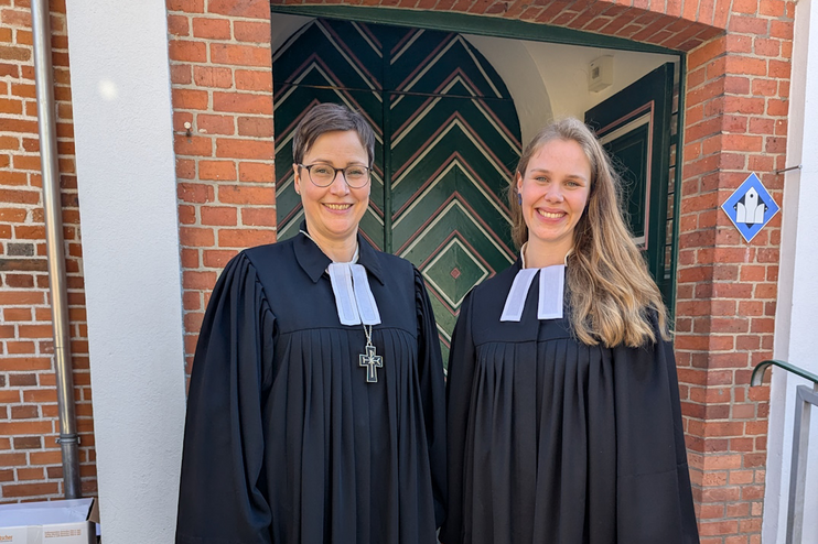 Zwei weiblich gelesene Personen in Talaren stehen vor einer Kirchentür.