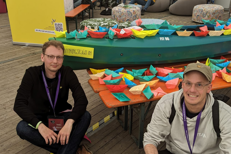 Zwei männlich gelesene Personen sitzen auf dem Holzfußboden in einem großen Zelt. Im Hintergrund ein Tisch mit Dutzenden Papierschiffen.