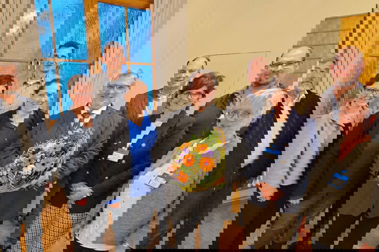 Eine Gruppe von als Frauen und Männer lesbare Personen, eine als Mann lesbare Person hält einen Blumenstrauß.