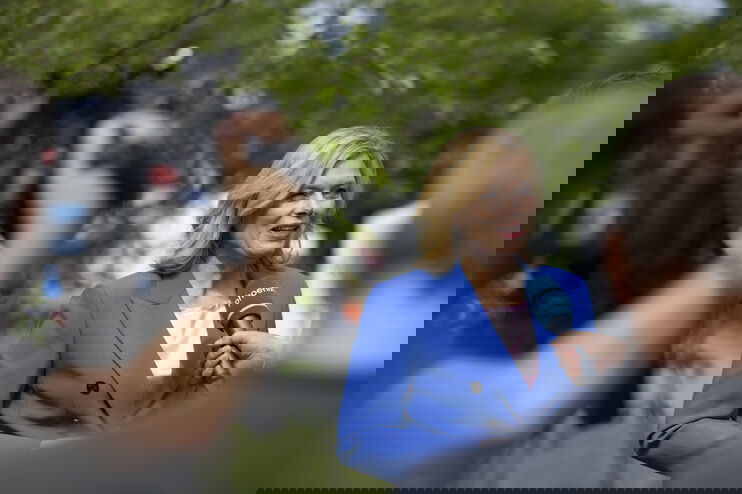 Im Vordergrund ist eine Kamera. Im Hintergrund gibt eine Frau ein Interview