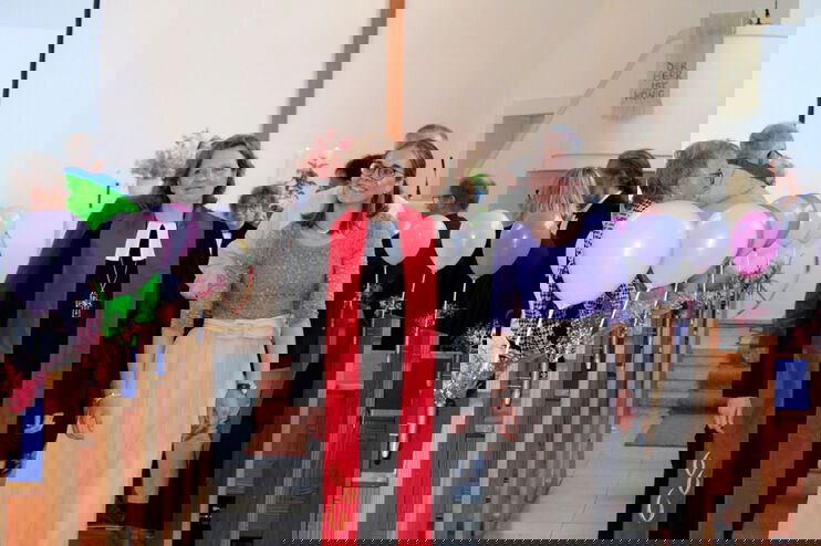 Zwei als Frauen lesbare Personen gehen durch den Mittelgang einer Kirche, links und rechts schweben Luftballons.