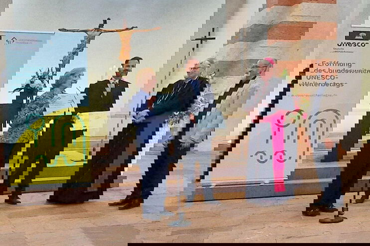 Eine als Frau lesbare Person und drei als Männer lesbare Personen stehen im Altarraum einer Kirche, zwei halten Schilder. Links steht ein Aufsteller mit der Aufschrift: Eröffnung des bundesweiten UNESCO-Welterbetags.