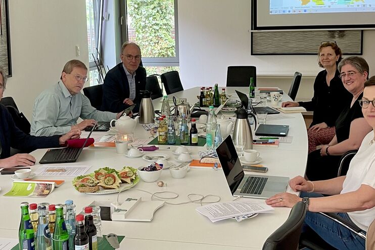 Eine Gruppe von sechs männlich bzw. weiblich gelesenen Personen sitzt an einem großen Konferenztisch; in der Mitte des Tisches liegen Arbeitsmaterialien sowie belegte Brötchen neben Getränken.