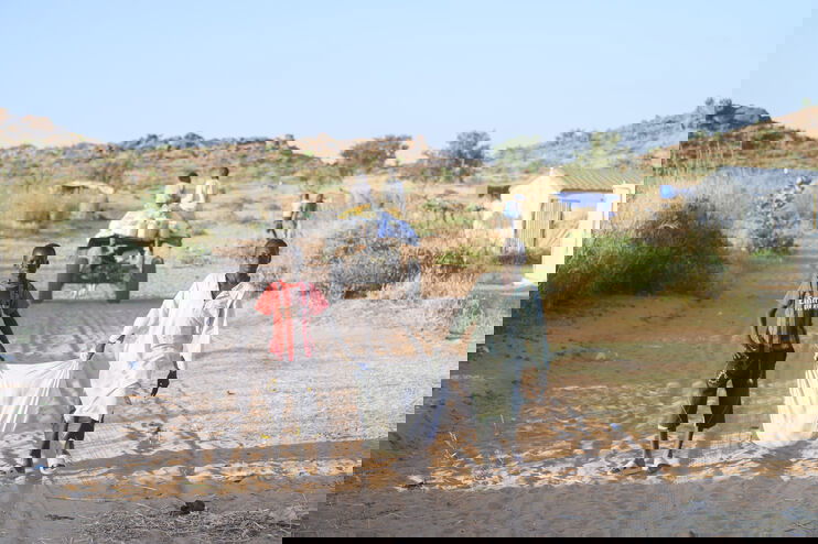 Zwei Menschen dunkler Hautfarbe laufen eine staubige Straße entlang und schleppen einen Sack zwischen sich. 