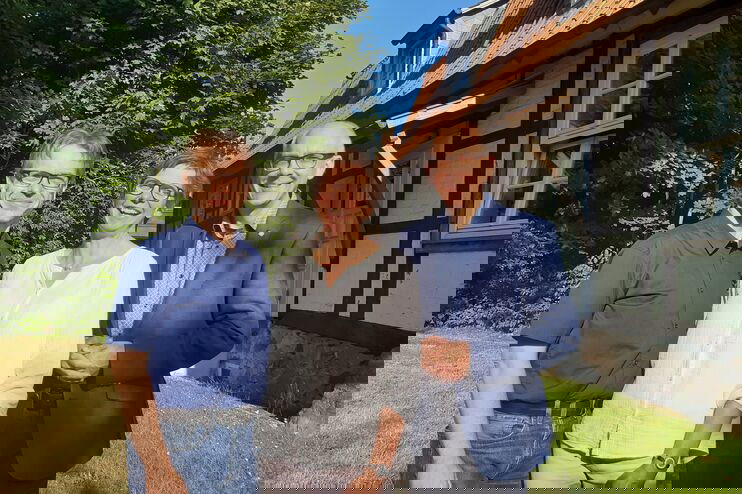 Zwei männlich und eine weiblich gelesene Person stehen vor einem Fachwerkhaus zusammen und lächeln in die Kamera.