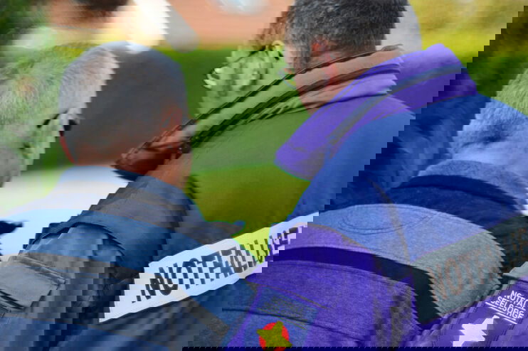 Zwei männlich gelesene Personen von hinten, einer trägt eine lila Jahre mit der Aufschrift: Notfallseelsorge