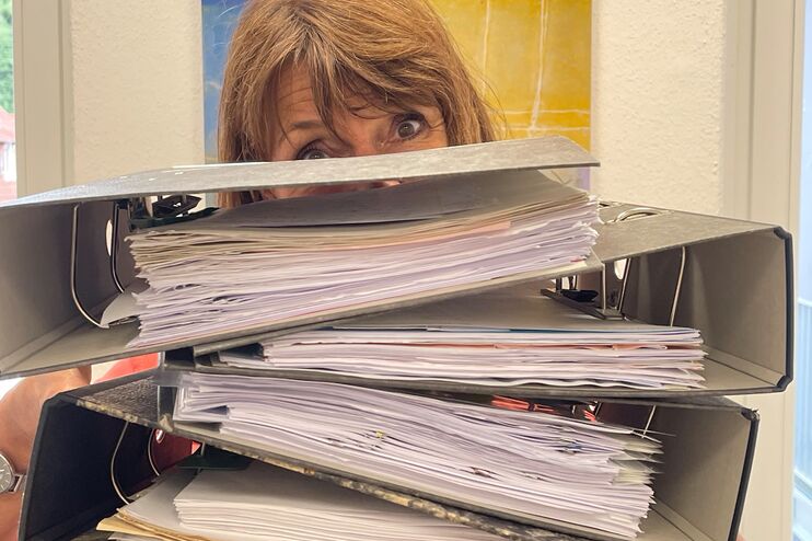 Eine weiblich gelesene Person, die hinter dicken Akten hervorschaut.