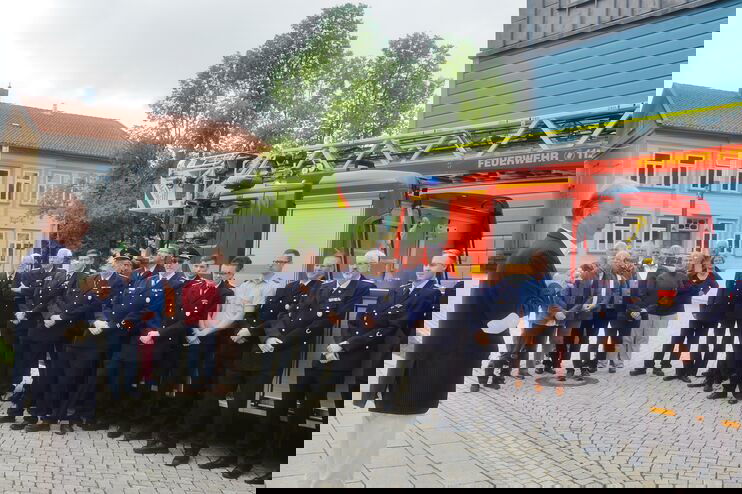 Mehrere Feuerwehrleute stehen vor einem Feuerwehrwagen und schauen eine männlich gelesene Person in heller Hose und dunklem Jackett an.