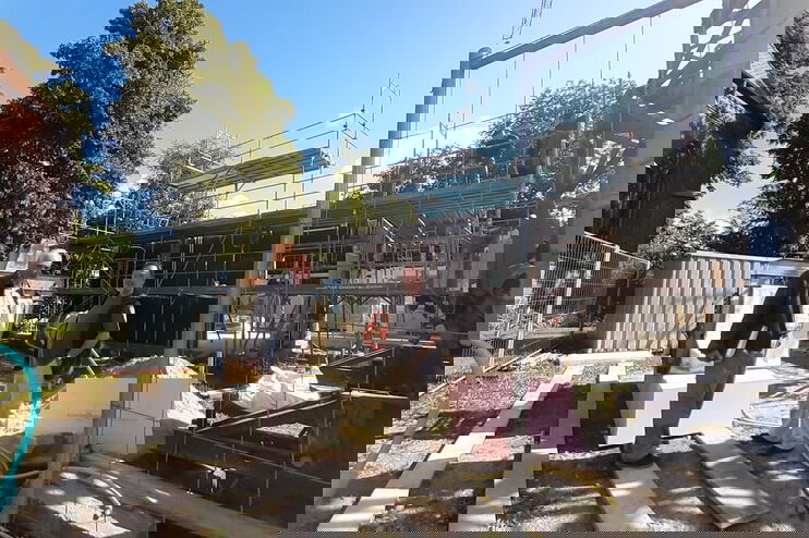 Mehrere Menschen stehen auf einer Baustelle, ein größeres Haus ist im Rohbau zwischen Bauzaun und Materialien zu erkennen.