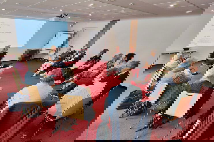 In einem Konferenzraum sitzen mehr als ein Dutzend Personen auf Stühlen in einem Kreis im Gespräch.