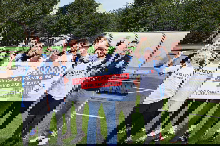 Eine Gruppe aus jungen Menschen steht auf einem Sportplatz und hält jubelnd einen symbolischen Scheck hoch.