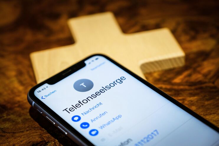 Ein Mobiltelefon, auf dessen Display "Telefonseelsorge" steht. Darunter liegt ein Holzkreuz.