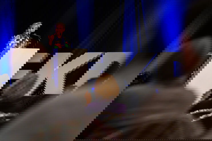 Eine als Frau lesbare Person steht auf einer Bühne am Redepult.