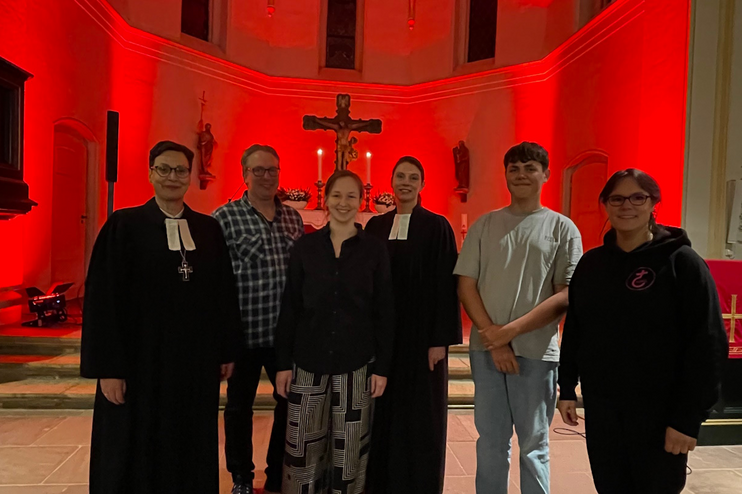 Sechs Personen, zwei im Talar, stehen in einer rot beleuchteten Kirche.