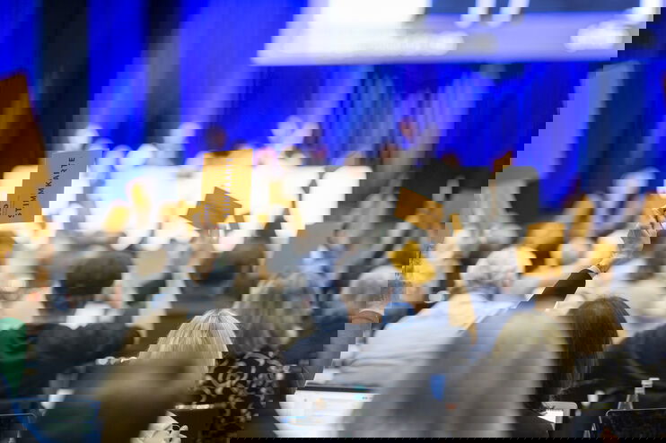 Menschen in einem blau beleuchteten Saal heben gelbe Karten zur Abstimmung.