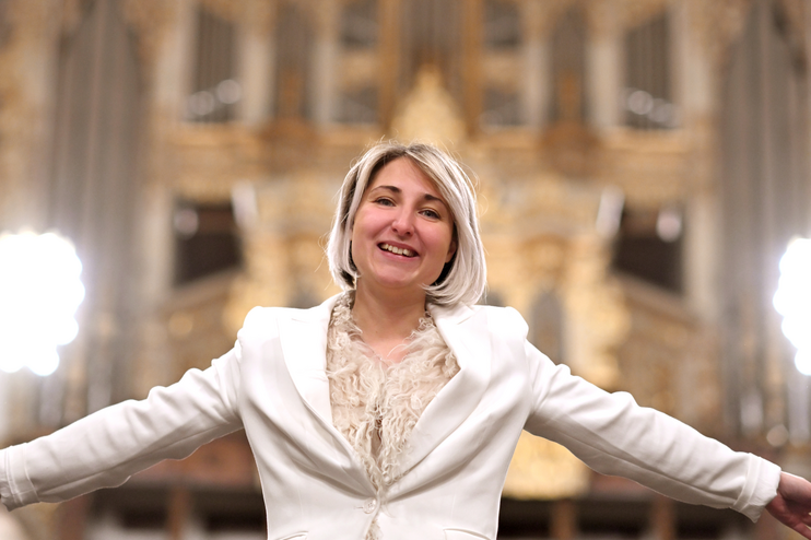 Eine weiblich gelesene Person in weißem Anzug strahlt vor einer goldenen Orgel.