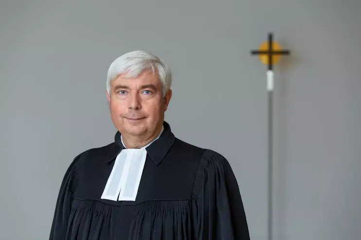 Eine als Mann lesbare Person mittleren Alters steht im Talar vor einem Kreuz.