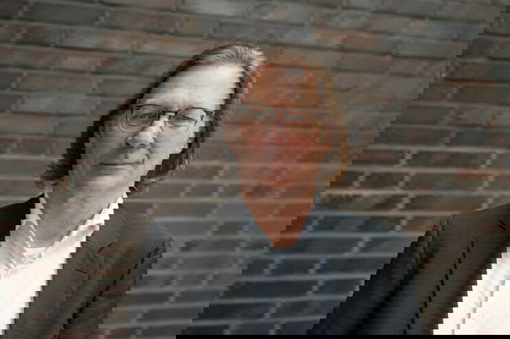 Eine männlich gelesene Person mit mittellangen Haaren, Brille, weißem Hemd und dunklem Sakko.