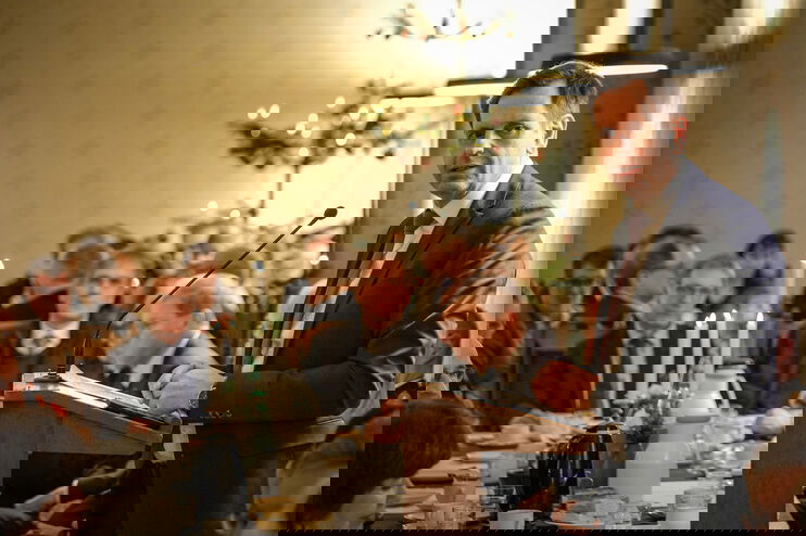 Eine männlich zu lesende Person steht an einem Redepult und spricht zu Menschen, die an einem festlich gedeckten Tisch sitzen.