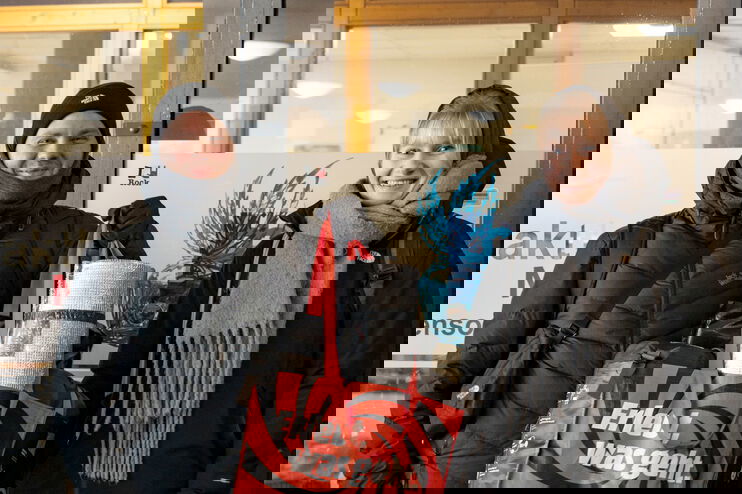 Eine als Mann und eine als Frau lesbare Person in Winterkleidung mit einem Beutel mit Schlafsack und Isomatte.