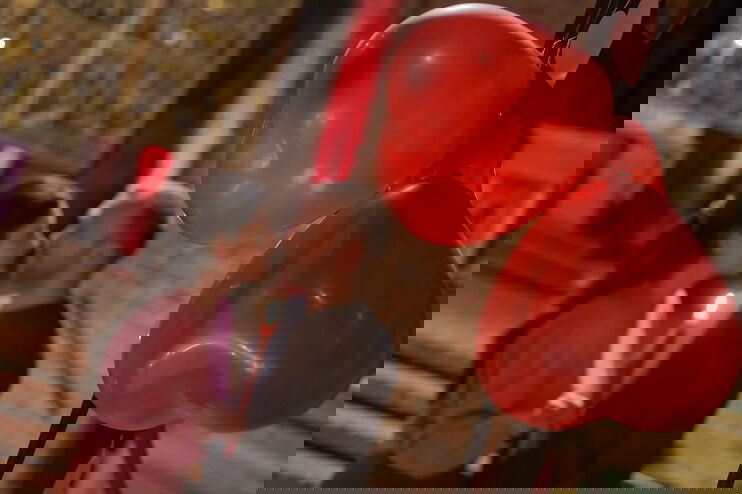 Zwei unscharf erkennbare Personen stehen sich eng gegenüber, eine Kerze in der Hand. Im Vordergrund sind Herz-Ballons scharf zu sehen.