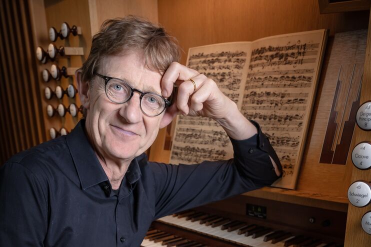 Eine männlich gelesene Person in blauem Hemd stützt sich mit einem Arm an einer Orgel ab.