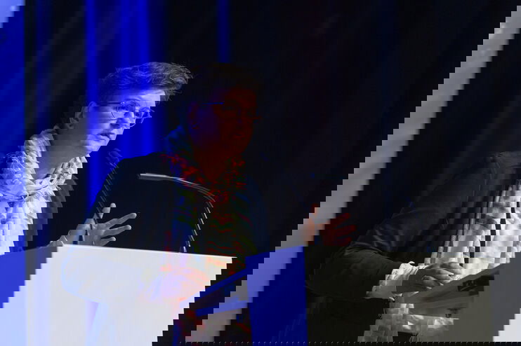 Eine als Frau lesbare Person spricht an einem Redepult vor einem blau angestrahlten Vorhang.