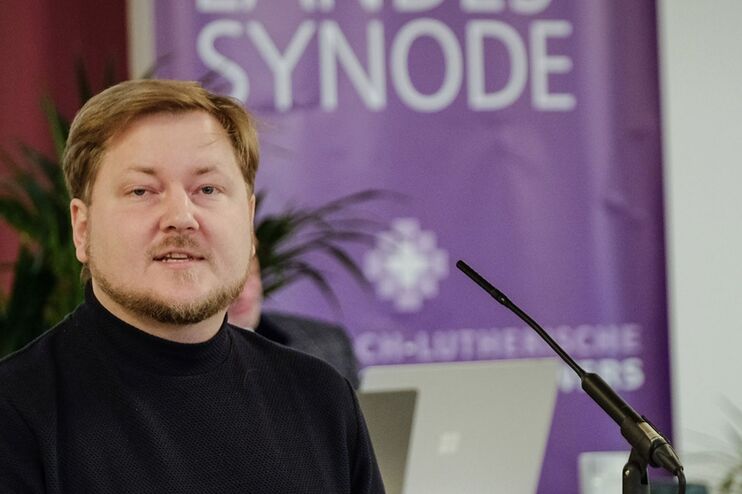 Ein Mann in einem schwarzen Pullover steht hinter einem Rednerpult, im Hintergrund hängt eine lila Flagge, auf der Landessynode steht.