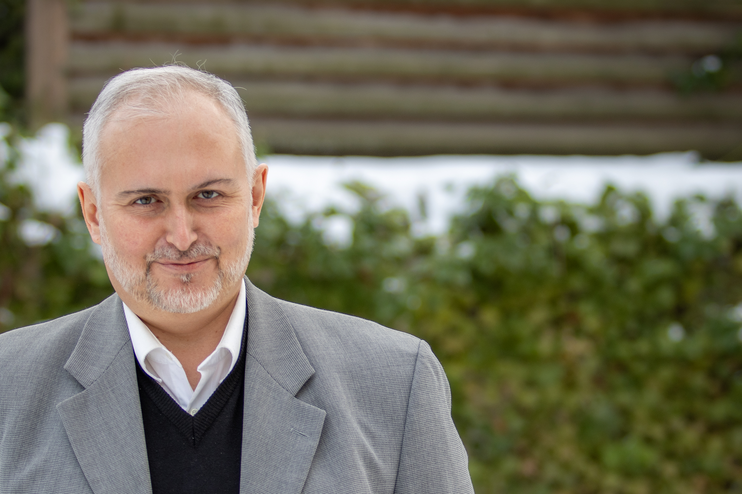 Eine männlich gelesene Person in grauem Jackett vor einer Hecke.