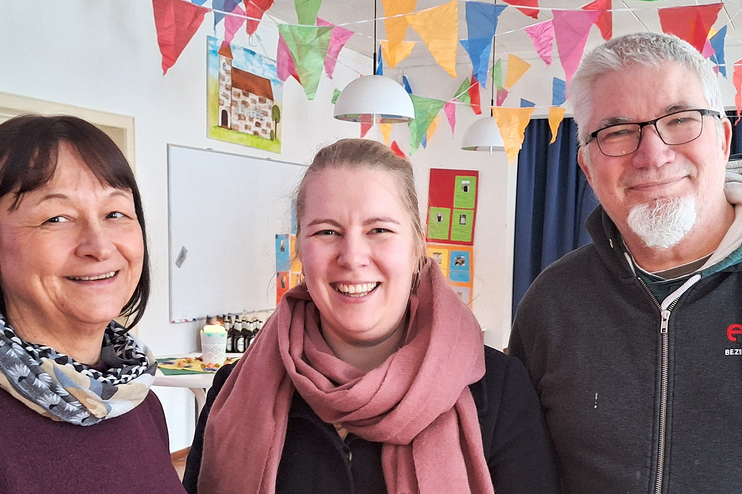 Drei Personen stehen lächelnd zusammen in einem bunt geschmückten Raum.