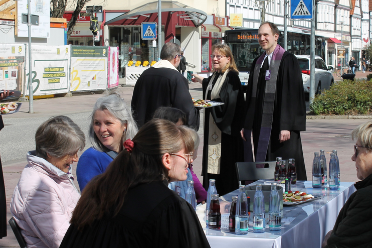 Mehrere Personen sitzen an einem gedeckten Tisch im Freien, einige Personen, teils in Talaren stehen darum.