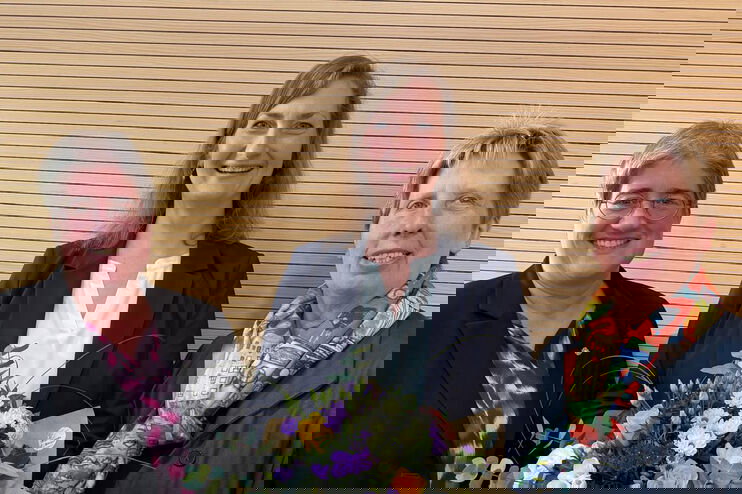 Drei als Frauen lesbare Personen, die mittlere hält einen Blumenstrauß.