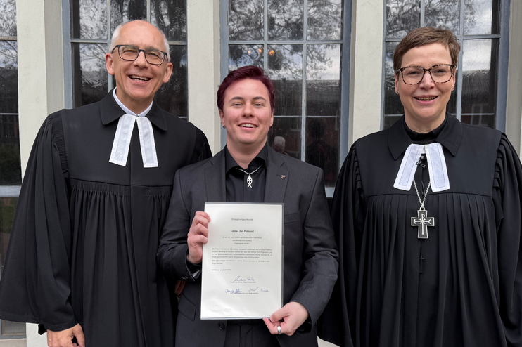 Drei Personen, zwei in Talaren, stehen zusammen, die dritte hält eine Urkunde in Händen.