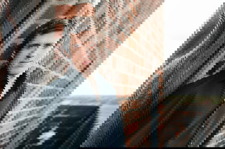 Eine als männlich lesbare Person im schwarzen Hemd lehnt an einer Backstein-Hauswand.