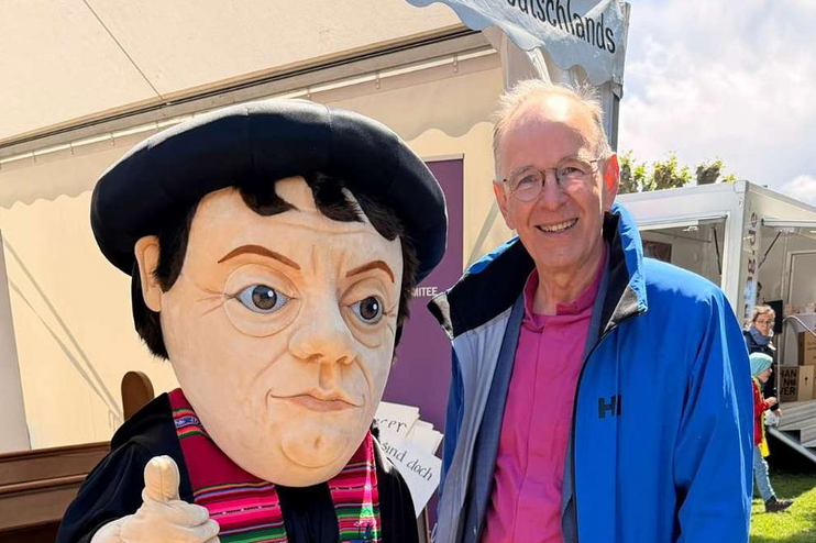 Eine männlich gelesene Person in blauer Jacke steht vor einem Zelt im Freien neben einer Figur, die Martin Luther ähnlich sieht. 