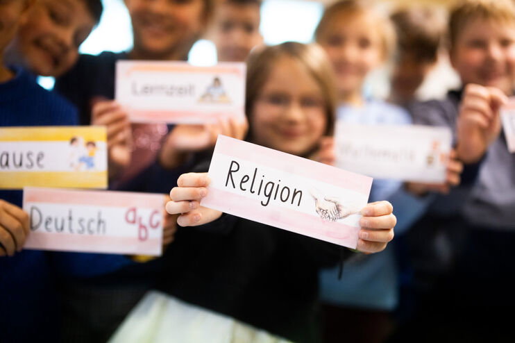 Kinder halten Schilder mit Schulfächern in die Kamera. Im Mittelpunkt steht Religion.