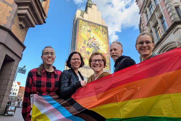 Mehrere als Männer und Frauen lesbare Personen mit einer Regenbogenflagge vor einem Kirchturm.