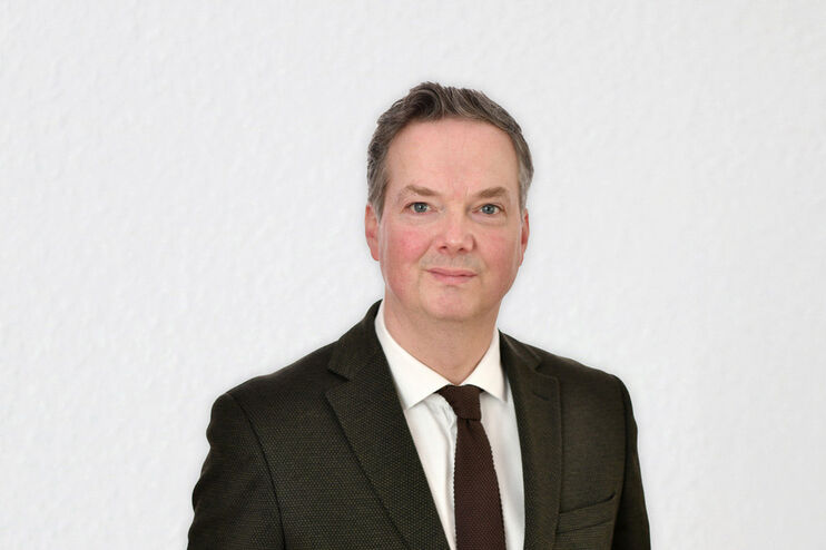 Eine als Mann lesbare Person mittleren Alters in Anzug und Krawatte vor einer weißen Wand.