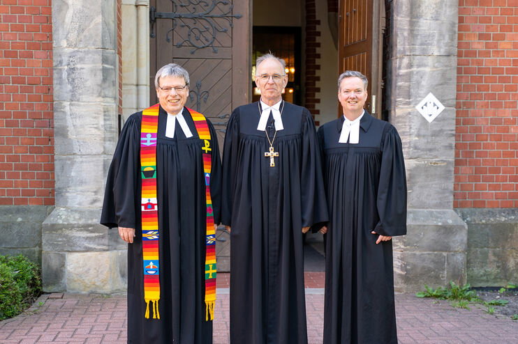 Drei als Männer lesbare Personen im Talar stehen vor der Einganstür einer Kirche.