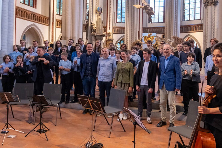 Mehrere als Frauen und Männer lesbare Personen stehen vor einem Orchester in einer Kirche.