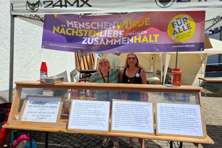 Zwei als Frauen lesbare Personen an einem Essenstand. Vorne in der Auslage liegt ein Statement zur Demokratie.
