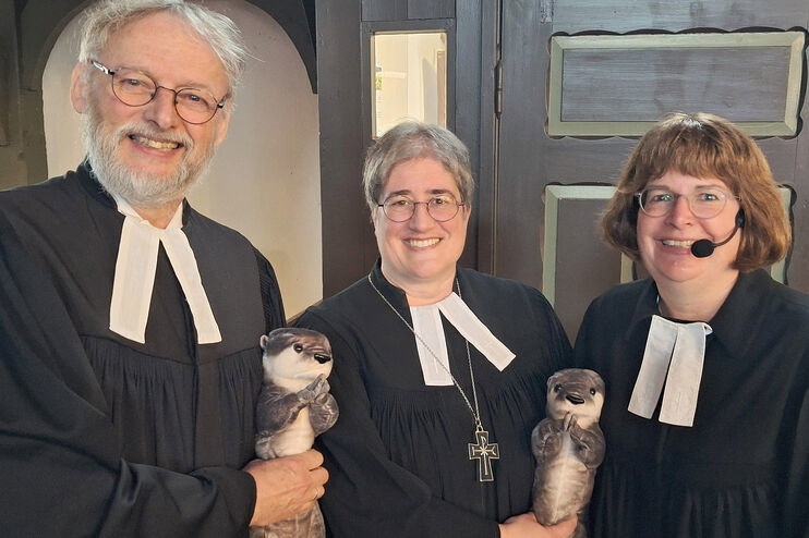 Zwei als Frauen und eine als Mann lesbare Personen im Talar halten einen Plüsch-Otter.