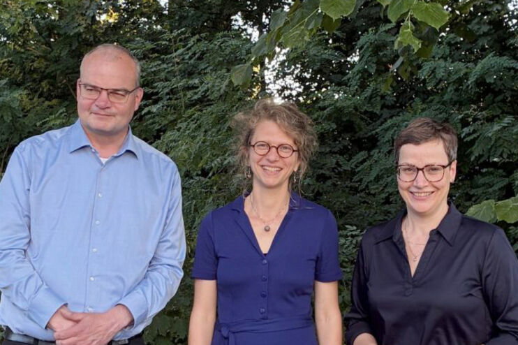 Zwei als Frauen lesbare Personen und eine als Mann lesbare Person stehen für ein Gruppenfoto vor Bäumen.