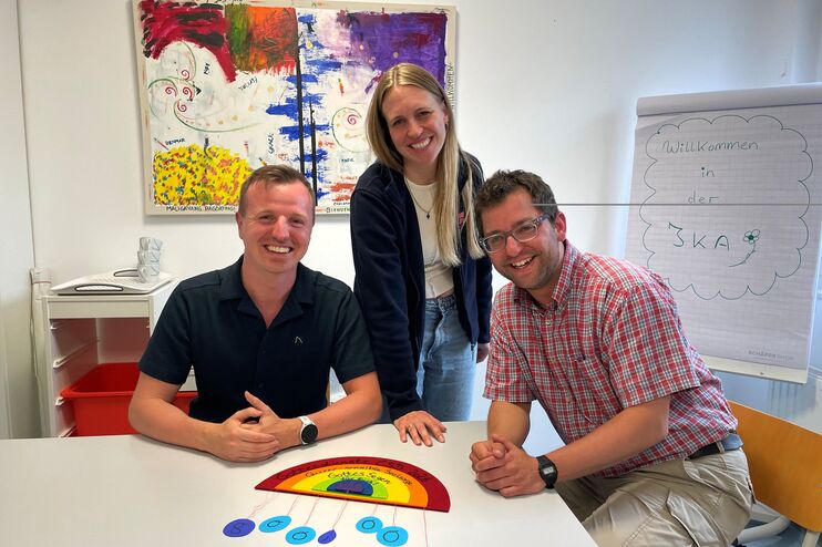 Drei lächelnde Personen sitzen an einem Tisch. Auf diesem Tisch liegt ein stilisierter Regenbogen. Im Hintergrund ist ein buntes Bild und eine Flipchart