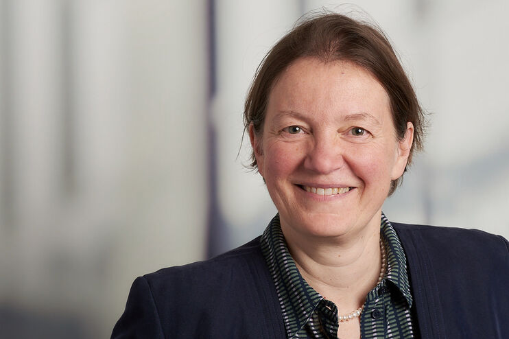 Portraitfoto einer als Frau lesbaren Person mit braunen zurückgebundenen Haaren.